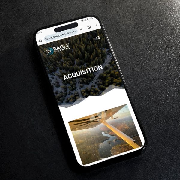 Smartphone on a dark surface displaying a webpage from Eagle Mapping. The screen shows the word “Acquisition” over a forest aerial view, with another image of a plane's wing above a landscape.