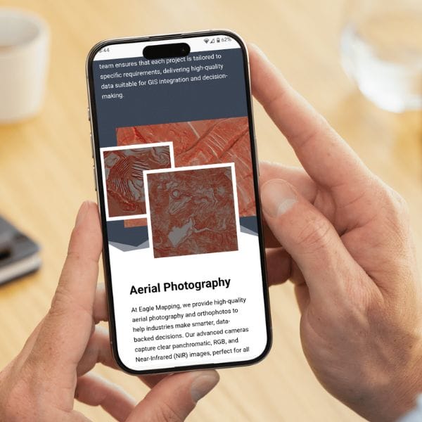 A person holding a smartphone displaying a webpage about aerial photography. The screen shows red-toned map images and text discussing high-quality data services for GIS integration and decision-making. A blurred desk and glass are in the background.