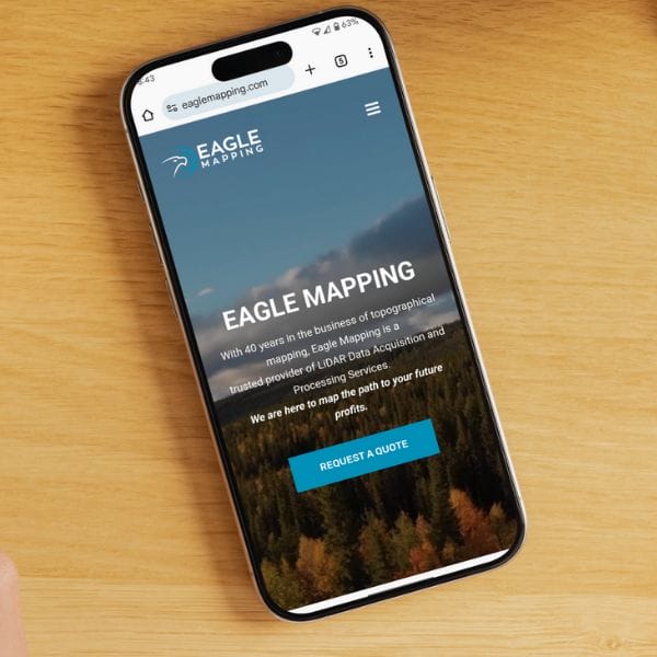 Smartphone on a wooden table displays the Eagle Mapping website. The screen shows a header image of a forest, company logo, and text highlighting their 40 years in topographical mapping. A blue button labeled "Request a Quote" is visible.