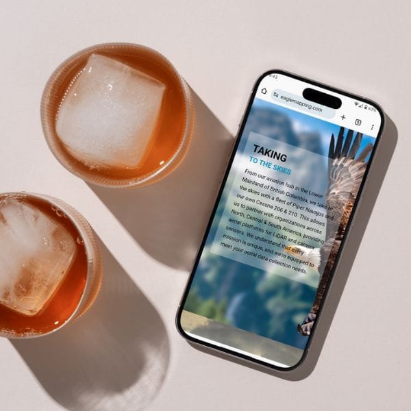 A smartphone displaying a webpage titled "Taking to the Skies" rests on a light surface. Two iced drinks in brown cups are beside the phone, with shadows cast by the sunlight.