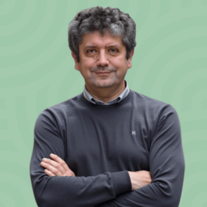 A person with curly hair and a slight smile stands with arms crossed, wearing a gray sweater over a collared shirt. The background is light green with a subtle circular pattern.