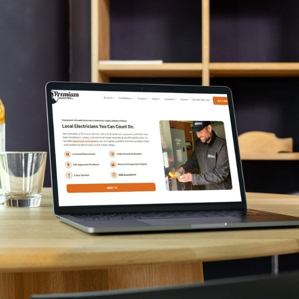 A laptop on a wooden table displays a website for electricians, featuring a man working and offering services like solar service and electrical repairs. A water glass and a dark blue chair are in the background.