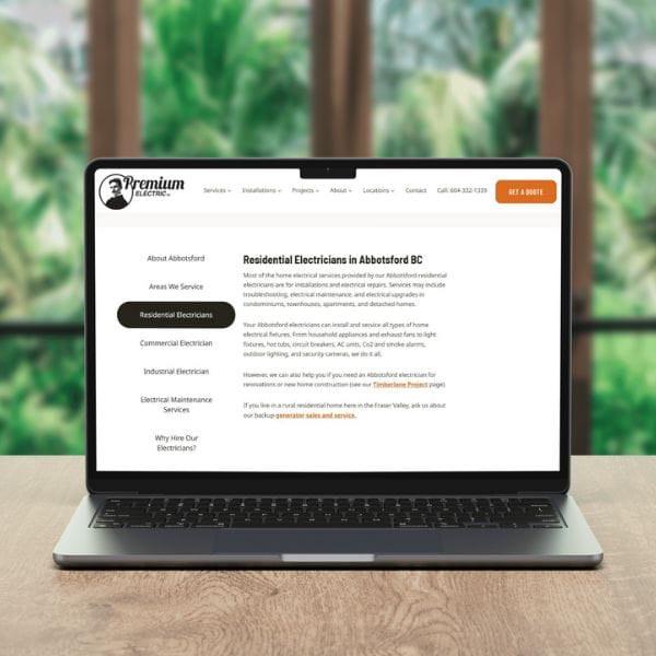 A laptop on a wooden table displays a webpage about "Residential Electricians in Abbotsford BC." The page has a sidebar with service categories and a blurred green background outside the window.