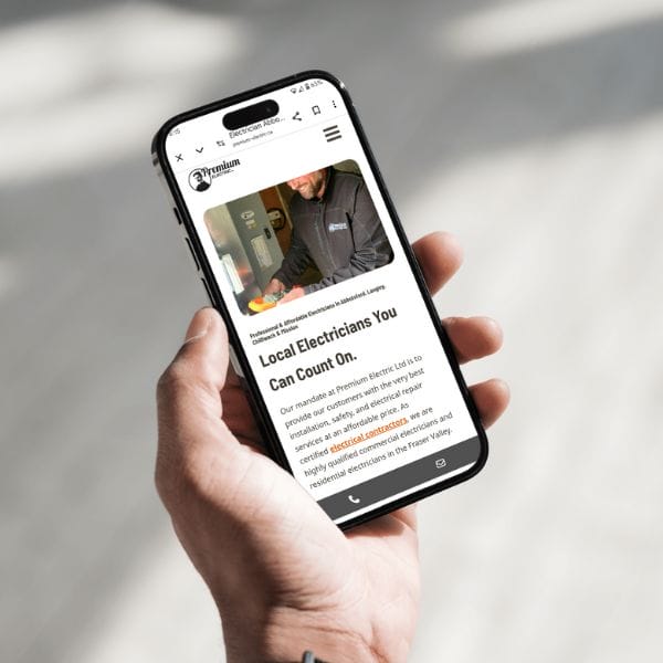 Person holding a smartphone displaying a website about local electricians. The screen shows a webpage with text and an image of a person in a dark jacket near electrical equipment.