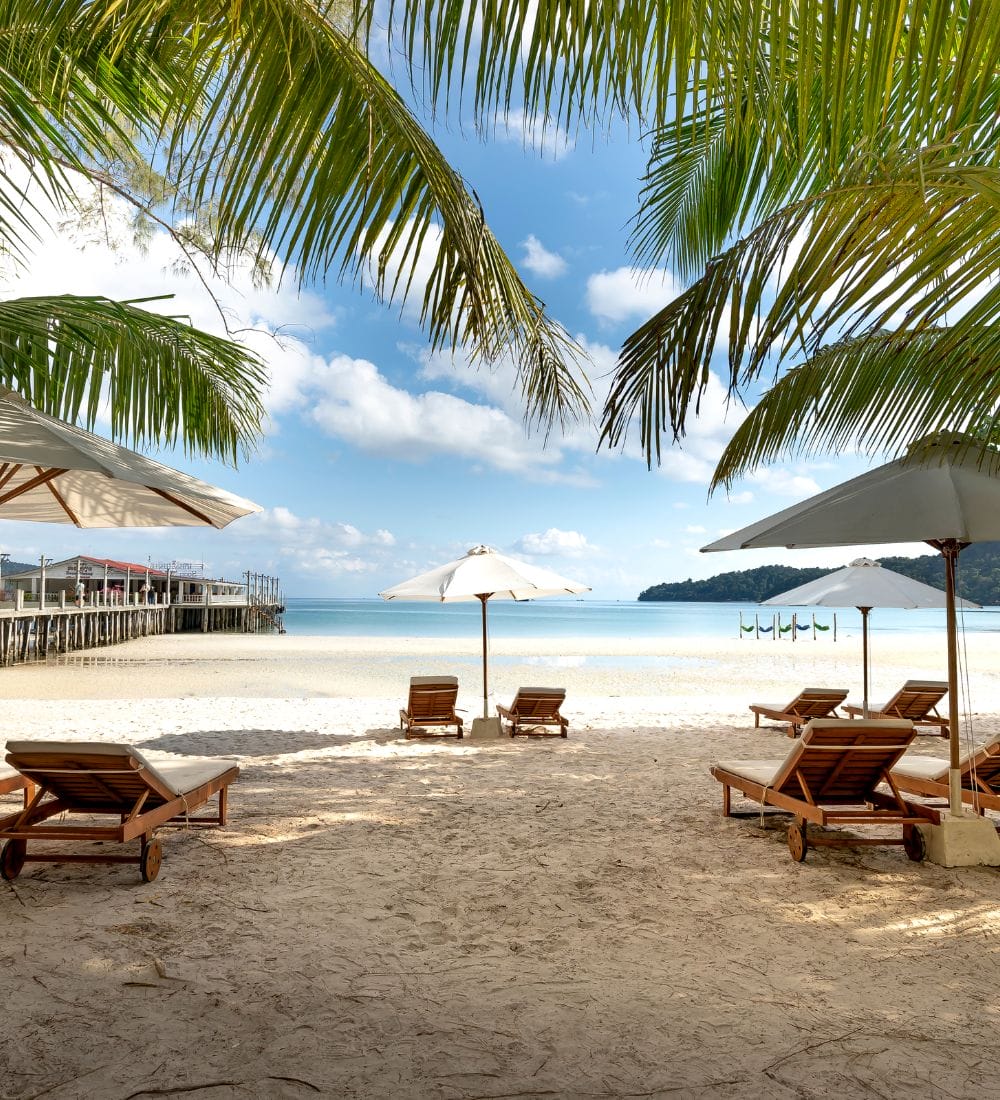 Sandy beach with wooden lounge chairs under white umbrellas, framed by palm leaves. A calm sea and a clear blue sky with scattered clouds are in the background. A pier extends into the water on the left side of the image.