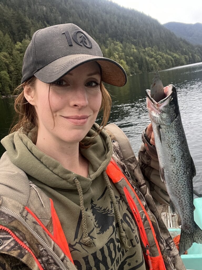 A person wearing a camo jacket and hat holds a fish on a boat. The background shows a lake surrounded by forested hills. The person is smiling, and the day is overcast.