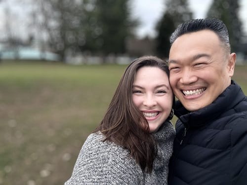 A smiling couple takes a selfie outdoors. The woman has long brown hair and is wearing a gray sweater, while the man has short dark hair and is wearing a black jacket. The background shows blurred trees and grass.