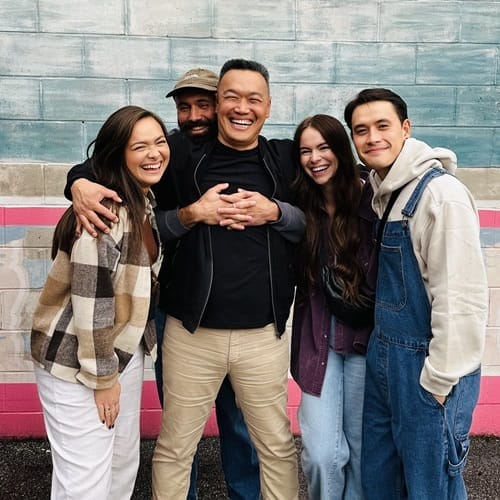 A group of five people standing closely together, smiling in front of a painted wall with blue and pink stripes. They are dressed in casual, warm clothing, and appear happy and relaxed.