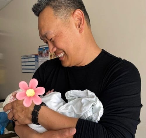 A smiling man in a black shirt cradles a newborn wrapped in a white blanket. The baby's face is covered with a pink flower emoji. The background shows a wall with charts and is softly lit.