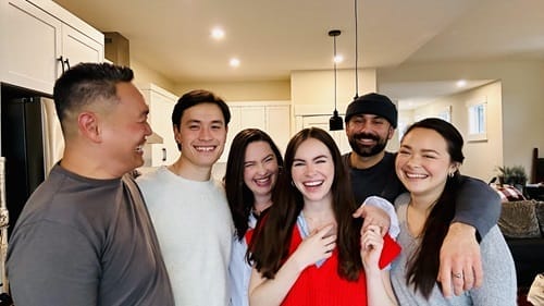 A group of six people stand closely together indoors, smiling and laughing. They are dressed casually and appear to be enjoying each other's company in a warmly lit room.