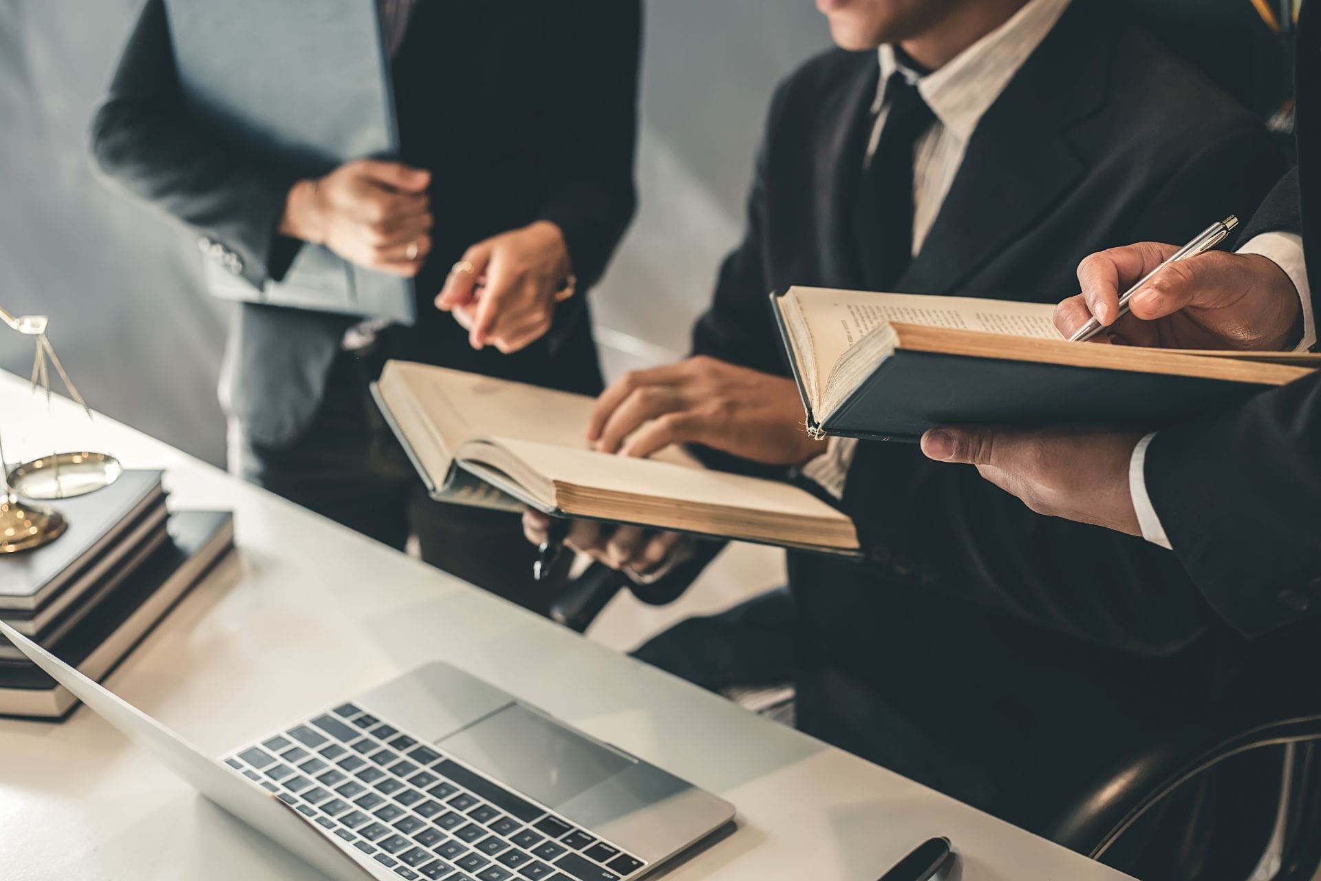Three people in business attire are gathered around a table with a laptop, holding and discussing open books. A set of scales and more books are on the table, suggesting a legal or professional meeting.
