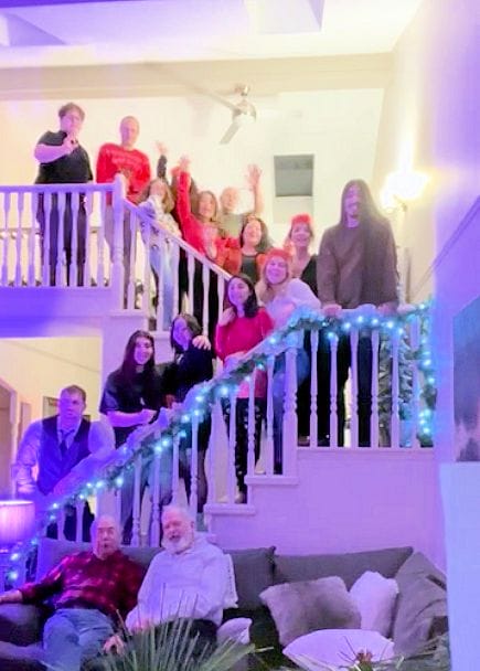 A group of people gathered on a staircase and a couch, smiling and waving at the camera. The staircase is decorated with blue and white lights, and a person stands at the top. The setting is indoors with warm lighting.