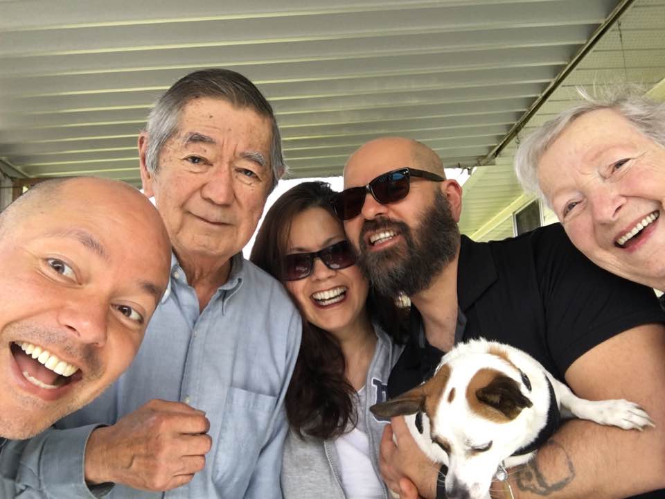 A group of five adults smiling under a covered porch, with one person holding a small dog. They appear happy and are closely gathered for the photo.