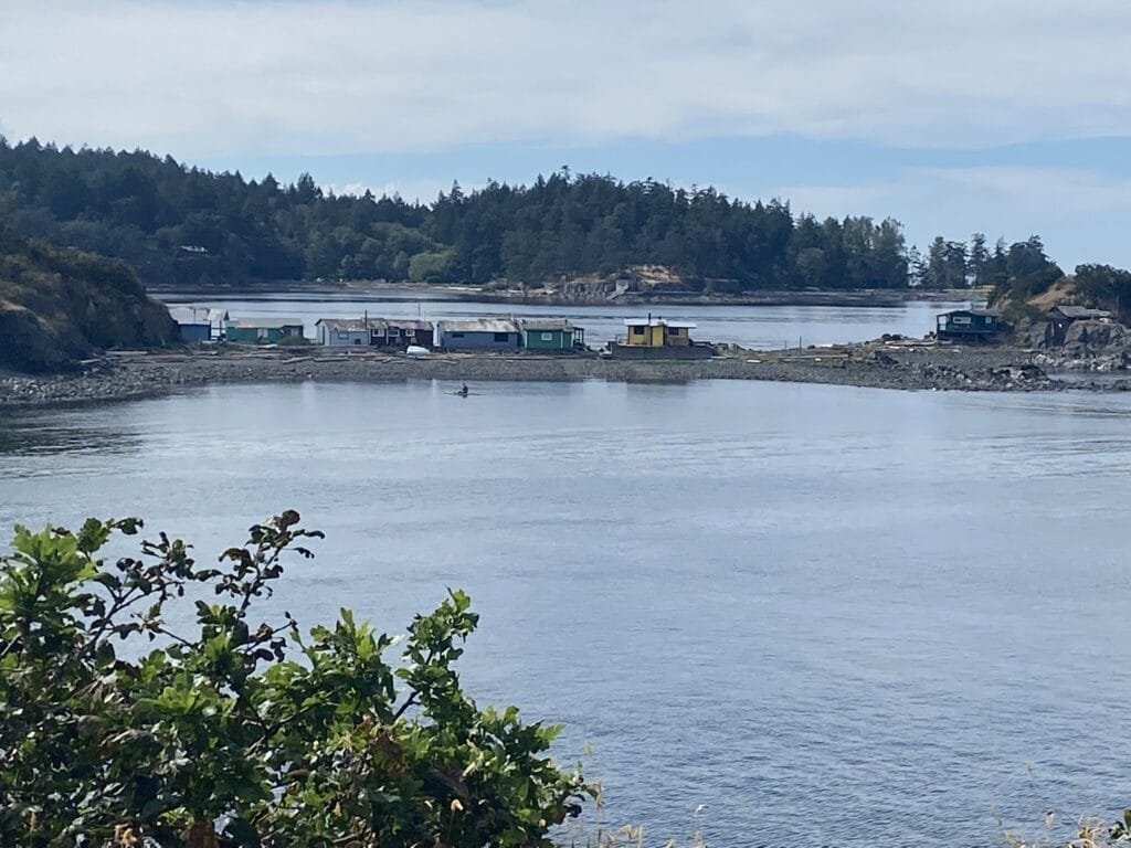 A calm waterfront scene with small colorful buildings along the shoreline, surrounded by green foliage and hills in the background. The water is still, reflecting the sky and trees.