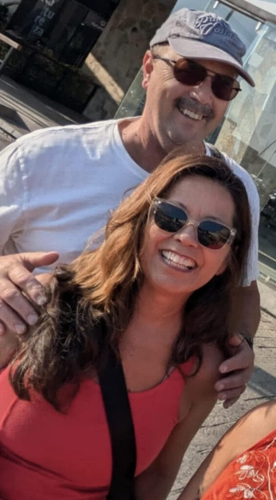 A man and woman are smiling outdoors. The man is wearing a cap, sunglasses, and a white shirt. The woman has long brown hair, sunglasses, and a coral top. They appear happy, and the setting is sunny.