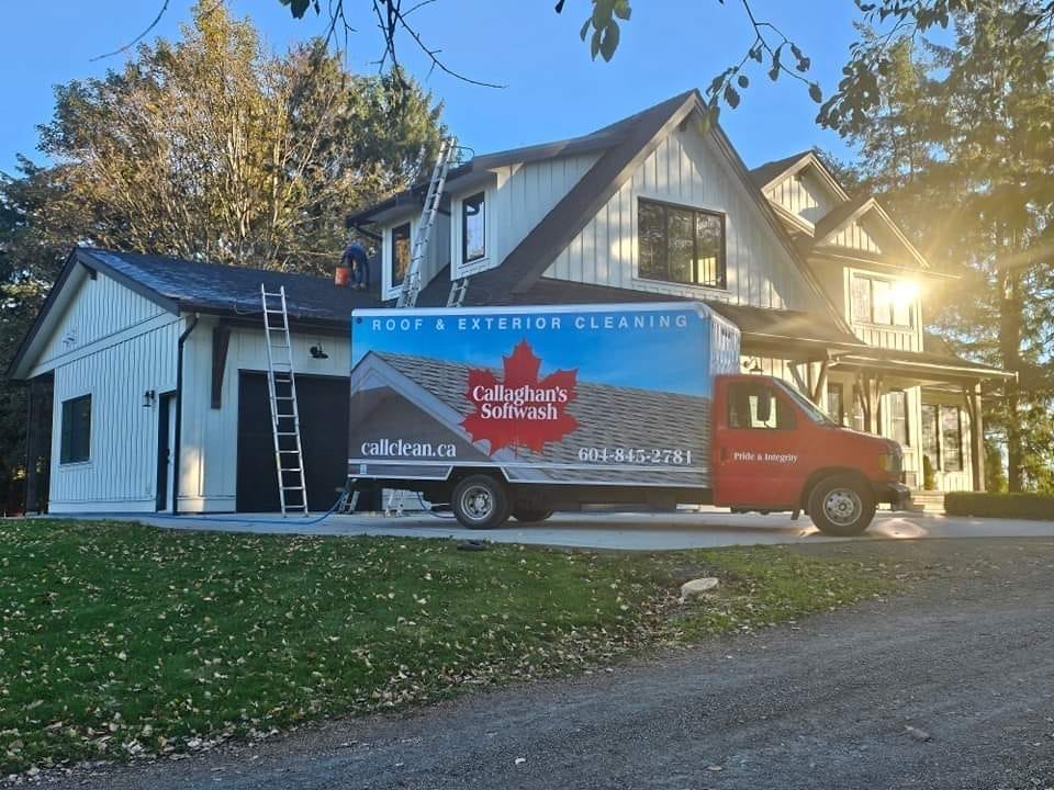 A red Callaghan's Softwash truck is parked beside a two-story white house with a black roof. A person stands on a ladder leaning against the house. The scene is lit by sunlight filtering through trees.