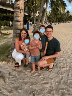 A family poses on a sandy beach, with palm trees and buildings in the background. The woman is kneeling beside three children, and the man is crouching on the other side. Hearts cover the children's faces. They all appear happy.