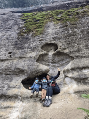 A person and two children sit in a natural rock formation with cavities, surrounded by textured stone and patches of moss. One of the children waves at the camera. They appear to be enjoying an outdoor activity.