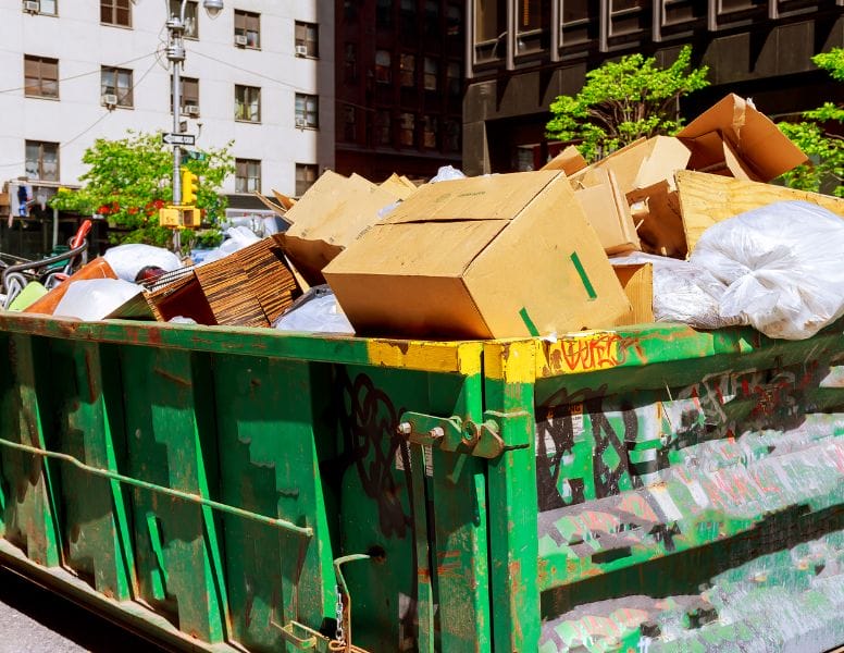 A large green dumpster on a city street overflowing with cardboard boxes, plastic bags, and assorted trash. Buildings and trees are visible in the background under a clear sky.