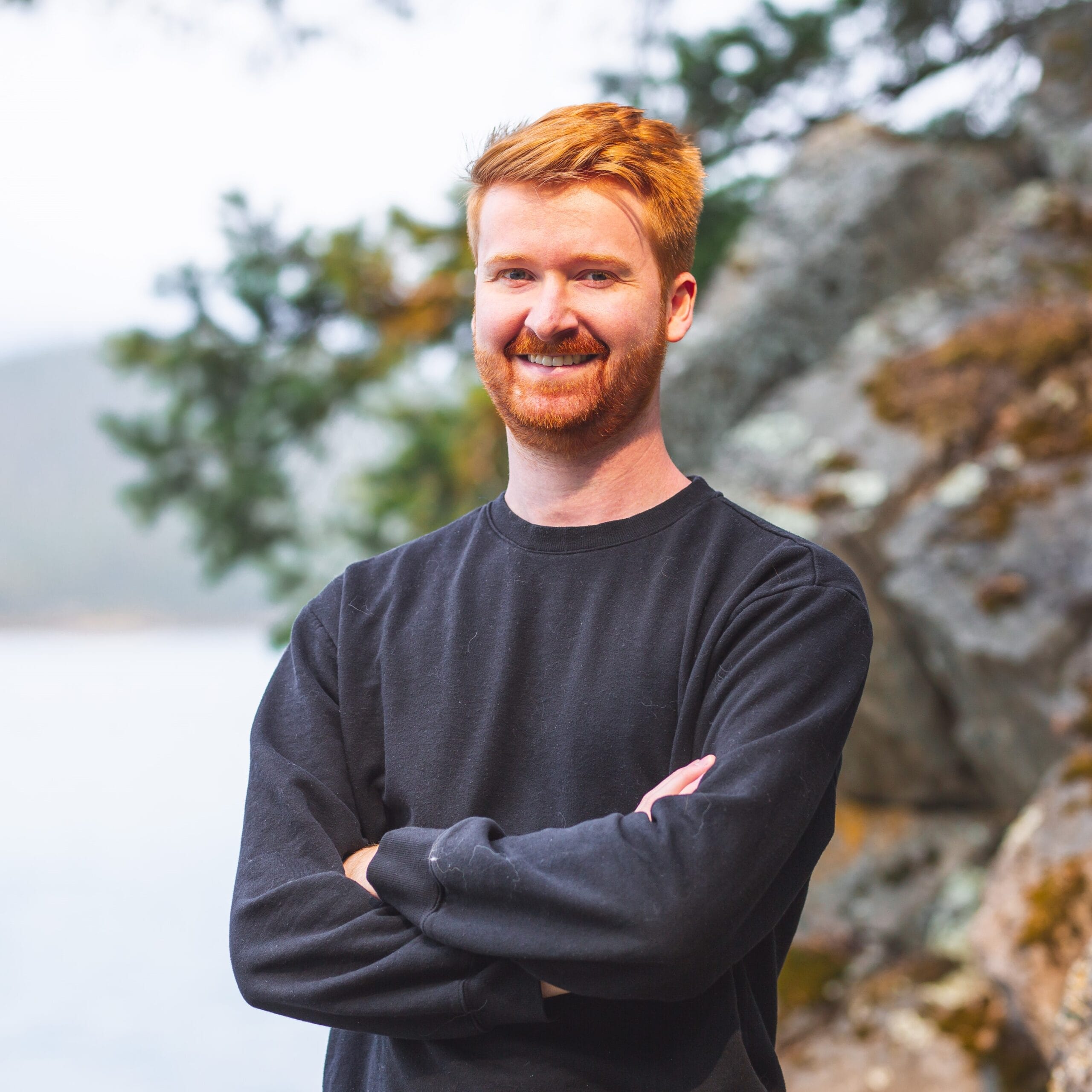 A person with red hair and a beard is smiling and standing with arms crossed, wearing a black sweater. The background features rocks, trees, and an out-of-focus body of water.