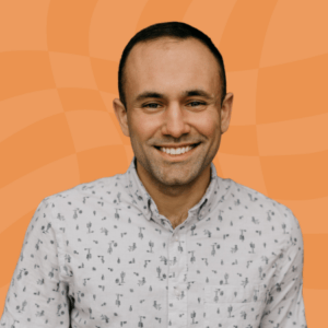 A person with short dark hair, smiling and wearing a light-colored button-up shirt with small patterns, stands in front of an orange wavy background.