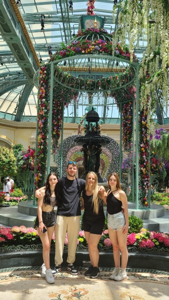 Four people stand arm-in-arm in front of a decorative floral fountain inside a greenhouse. The fountain is adorned with colorful flowers, and the sunny setting is filled with greenery. They all smile for the photo.