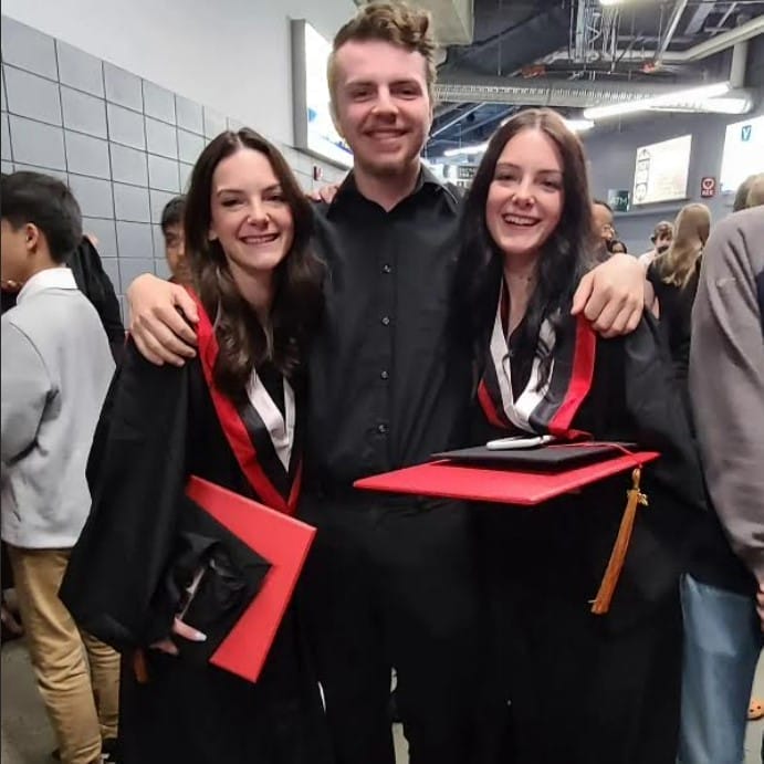 Three people stand together in a crowded indoor setting. Two are wearing black graduation gowns and holding red diplomas, smiling. The third person is in the middle, wearing a black outfit. They appear happy and are embracing each other.