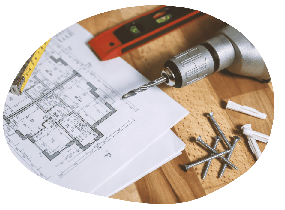 A drill, measuring tape, and spirit level are placed on a wooden surface alongside screws and wall plugs. Blueprints of a floor plan lay beneath them, suggesting a construction or renovation setting.