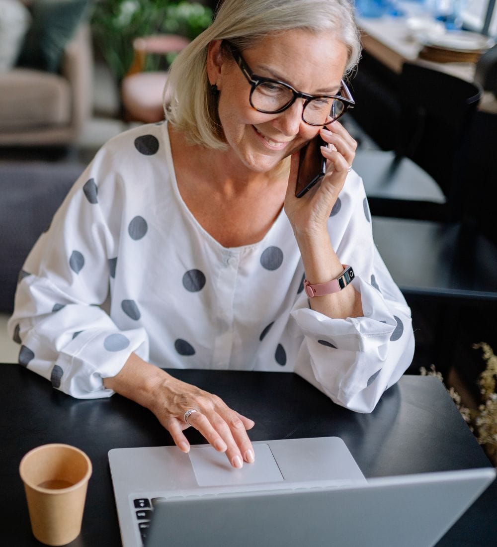 Older woman with glasses, wearing a white polka dot shirt, sits at a table while talking on a smartphone and using a laptop. A small coffee cup is beside her. The background is a cozy, blurred sitting area with plants.