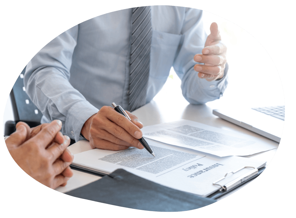 Two people in business attire sit at a desk, reviewing documents labeled "Insurance Policy." One person gestures while the other points at the papers with a pen; a laptop and calculator are also visible.