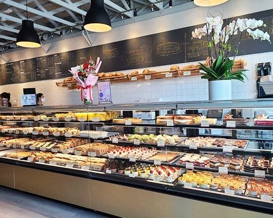 A bakery display filled with assorted pastries, cakes, and desserts. Behind the glass counter, shelves hold loaves of bread. Flowers and a potted plant decorate the counter, and a chalkboard menu is on the wall.