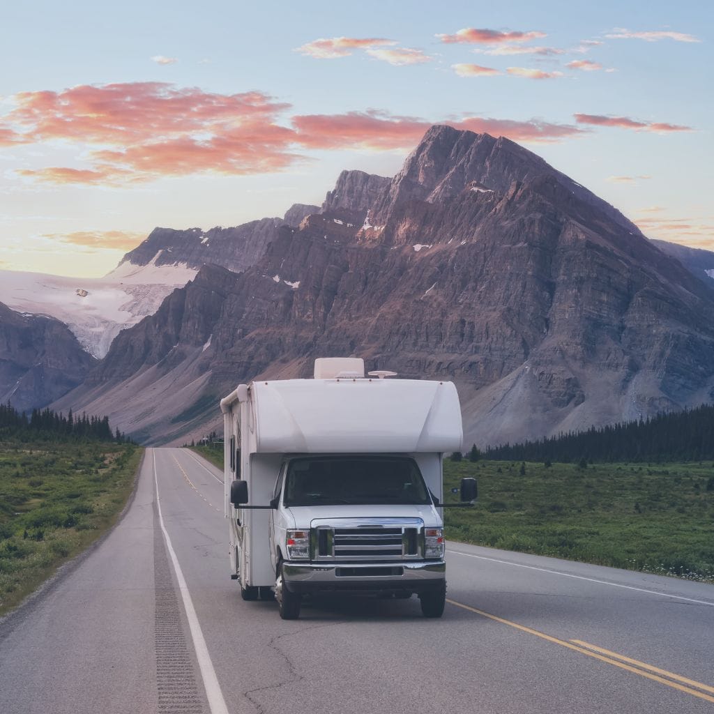 A white RV drives down an empty road surrounded by greenery, with dramatic mountains and a partly cloudy sky with pinkish sunset hues in the background.