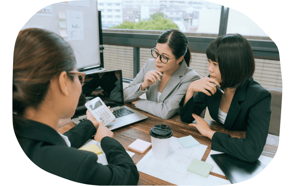 Three women in business attire sit around a wooden table with a laptop, documents, coffee cup, and sticky notes, discussing industry specific web design as they look at a calculator one of them is holding.